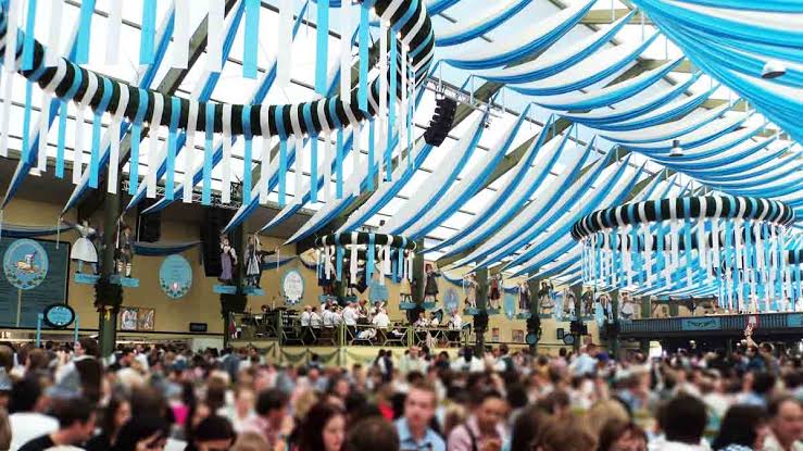 Một gia đình ngồi tại bàn dài trong lều bia, trẻ em đội mũ truyền thống Bavaria cười vui vẻ